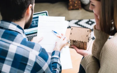 Two people sit together reviewing paperwork with a laptop; one holds a pen and the other holds a small model house, suggesting a discussion about home buying, real estate, or next steps after a car accident.