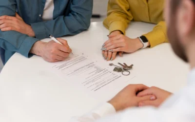 After a car accident, three people sit at a white table with documents and a set of keys; one person is signing papers while another observes.