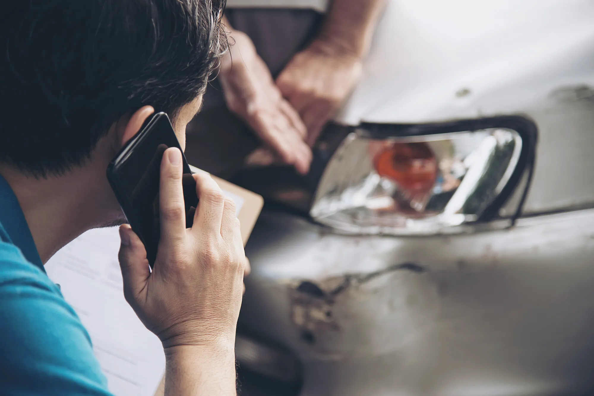 A person makes a phone call while another inspects damage to the front bumper of a silver car, likely documenting the aftermath of Motor Vehicle Accidents.