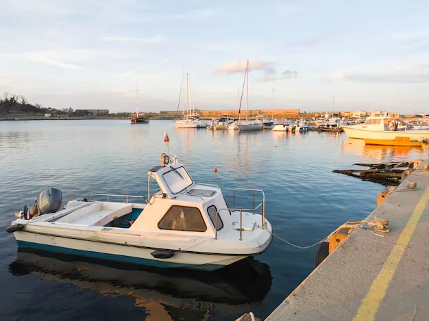 A small white motorboat is docked at a concrete pier in a calm harbor, highlighting the importance of safety to prevent motor vehicle accidents, with sailboats anchored in the background under a clear sky.