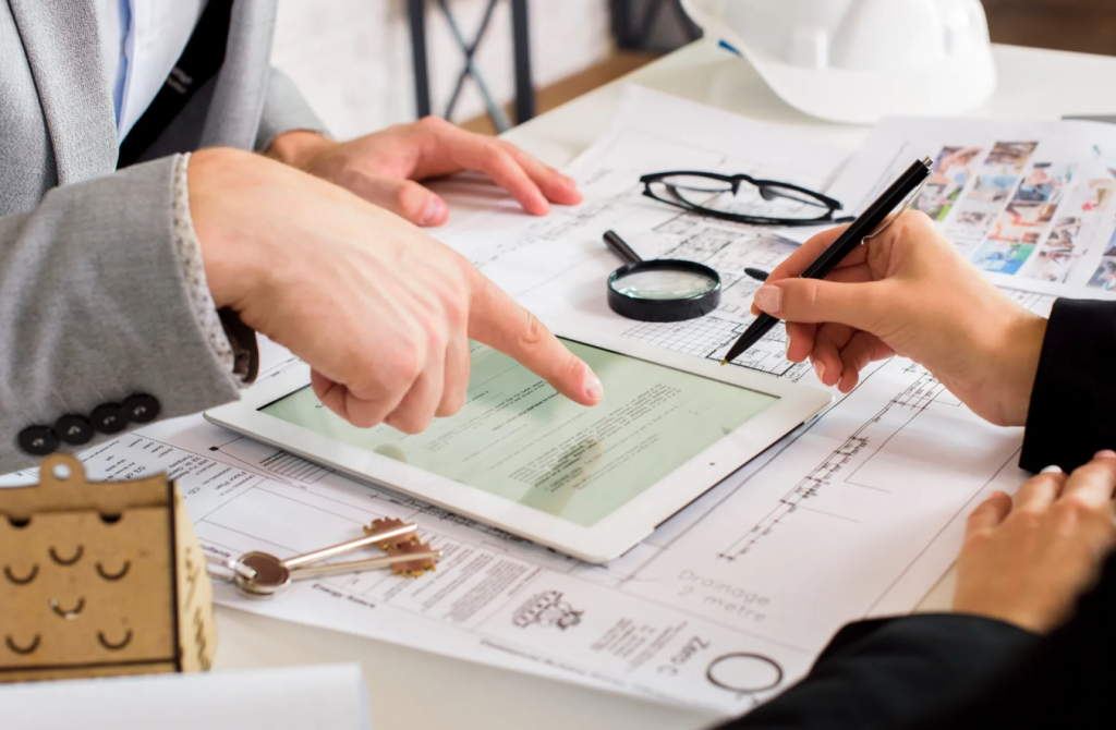 Two people review documents and blueprints on a table, exploring various practice areas with a tablet, pencils, and magnifying glass; a hard hat and keys are also visible.