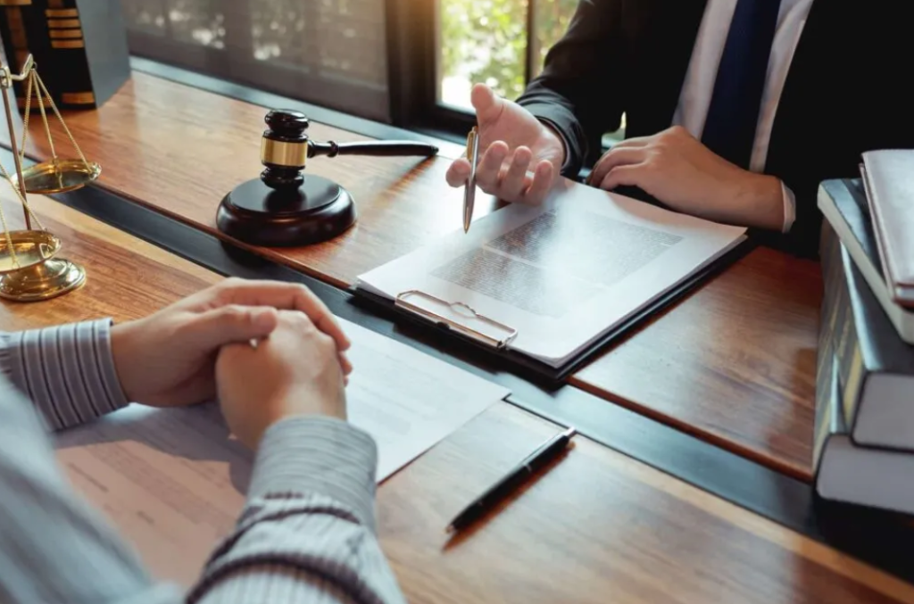 Two people sit across a desk with legal documents, a gavel, and scales of justice, discussing various practice areas in a professional legal setting.
