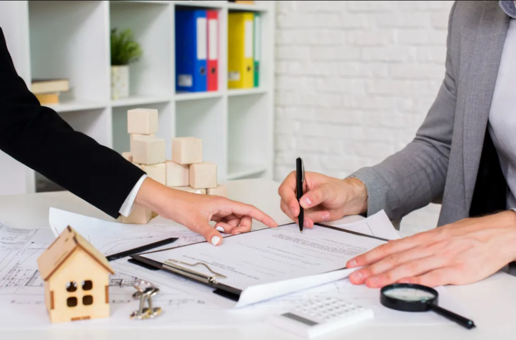 Two people sit at a desk reviewing and signing documents related to different practice areas. A miniature house, wooden blocks, magnifying glass, and office supplies are on the table. Shelves with folders fill the background.