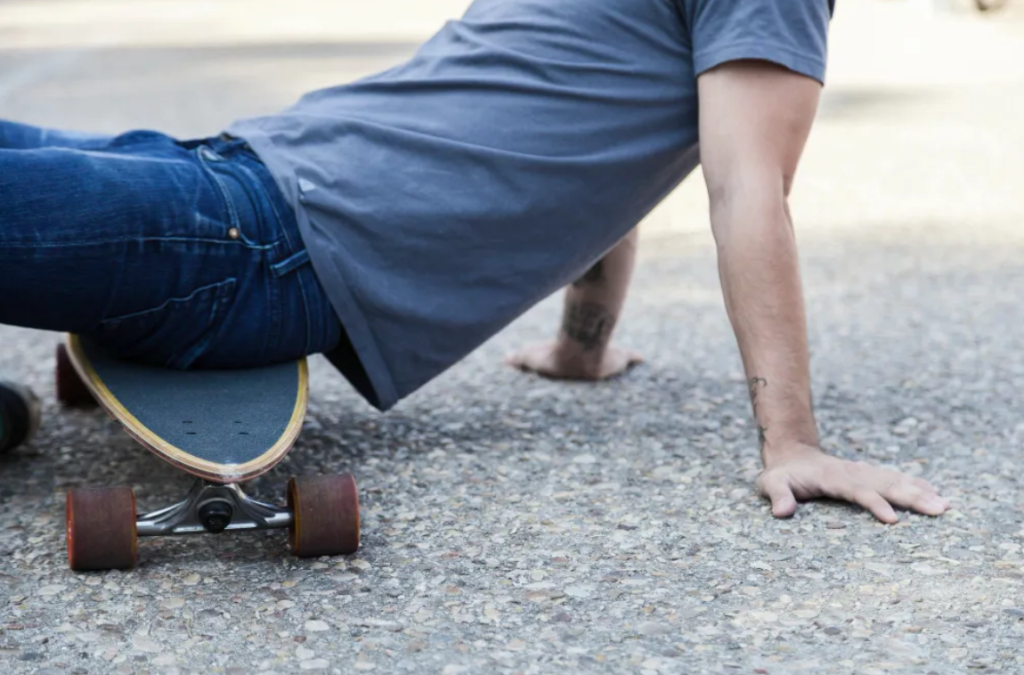 A person wearing jeans and a gray shirt sits on the ground with one hand on the pavement and a skateboard beneath them, appearing to recover from a slip and fall.