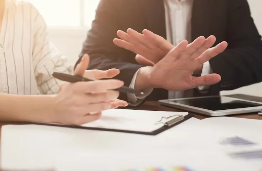 Two people sit at a table with documents and a tablet; one gestures with hands crossed in a stopping motion while discussing property damage, as the other holds a pen, ready to take notes.