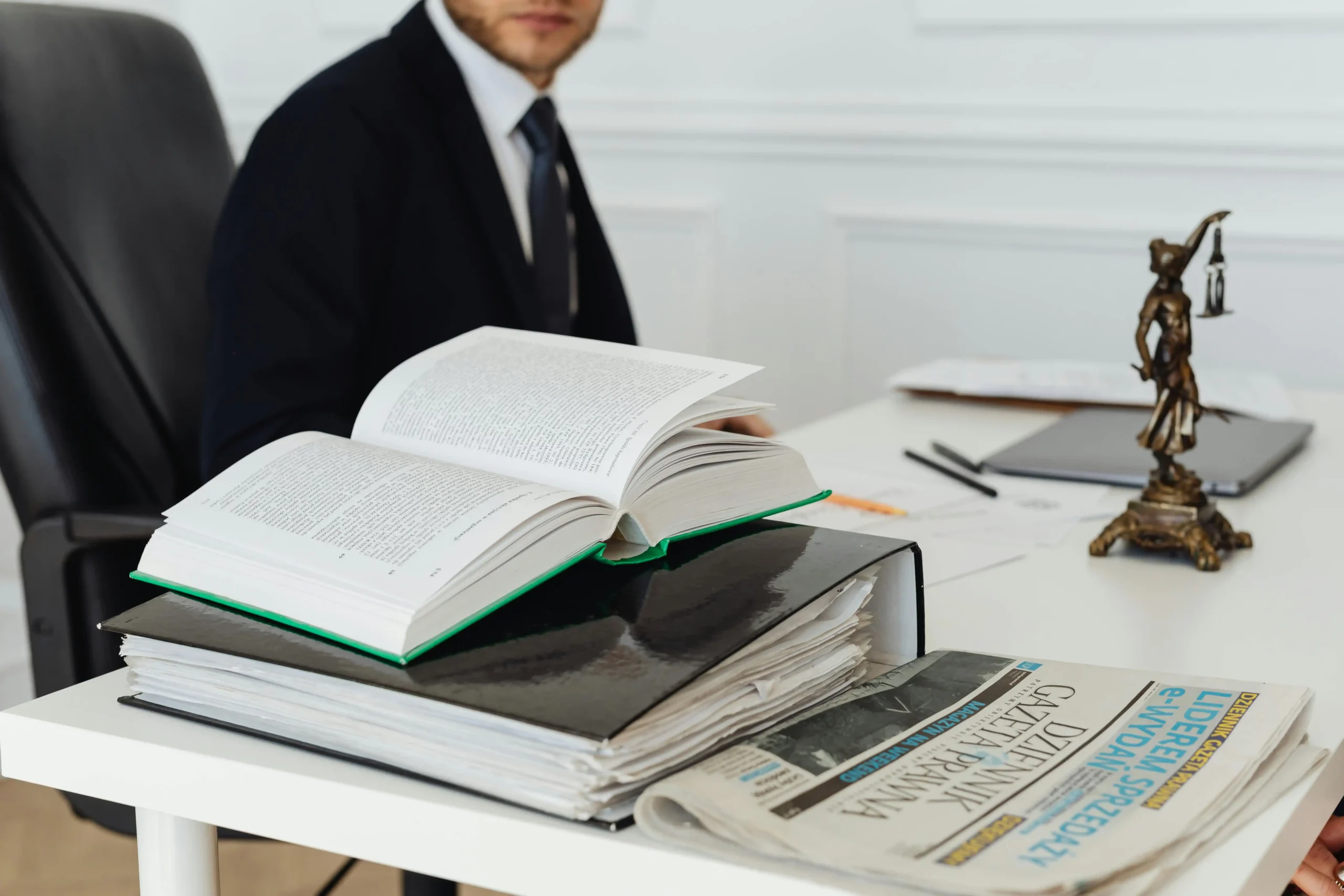 A person in a suit sits at a desk with open books, a stack of files, a newspaper, and a small Lady Justice statue, appearing to review documents related to Homeowner Association (HOA) disputes in an office setting.