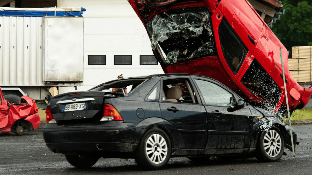 A red car crashed on top of a black car, with broken glass and visible damage, highlighting the aftermath of motor vehicle accidents in what appears to be a scrapyard or industrial area.
