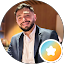 A man with a beard, wearing a suit and light-colored shirt, is smiling at the camera. There is a colorful circular badge with a white star in the bottom right corner.