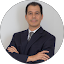Isaac Kassab, dressed in a dark suit and tie, stands with his arms crossed, facing the camera against a plain light background.