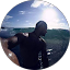 Person in a wetsuit stands near the ocean, looking at the water under a bright sky with scattered clouds.