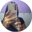 Person with long blue nails holds a smartphone in front of their face, taking a mirror selfie against a purple background.