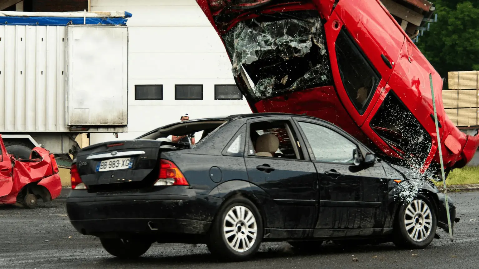 A red car crashed on top of a black car, with broken glass and visible damage, highlighting the aftermath of motor vehicle accidents in what appears to be a scrapyard or industrial area.